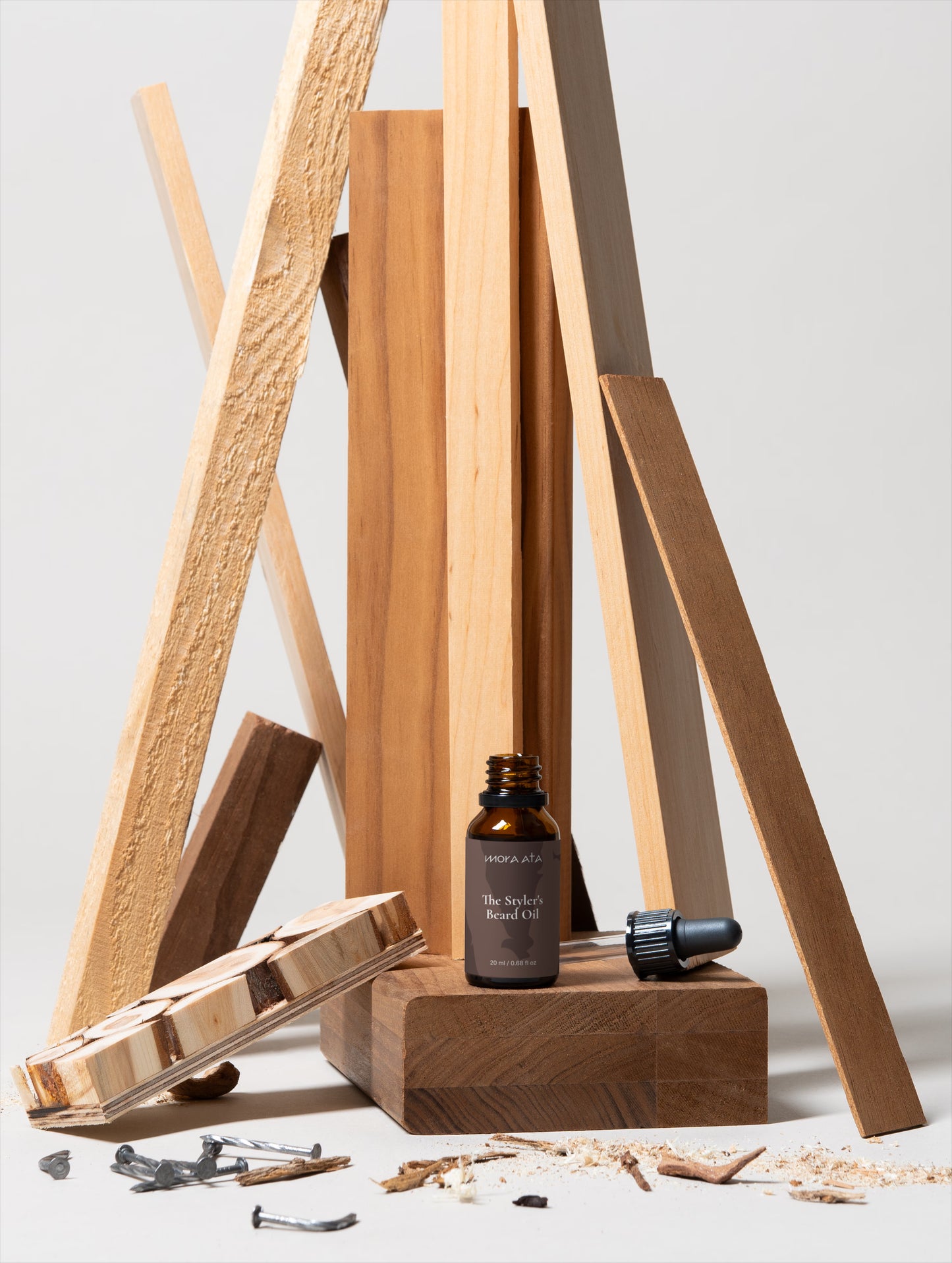 A bottle of Mora Ata The Styler's Beard Oil bottle and a dropper on a wooden stand with natural elements on a light background