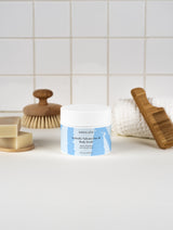 Jar of Mora Ata Icelandic Volcano Face & Body Scrub with a brush, soap, and towel on a tiled bathroom counter