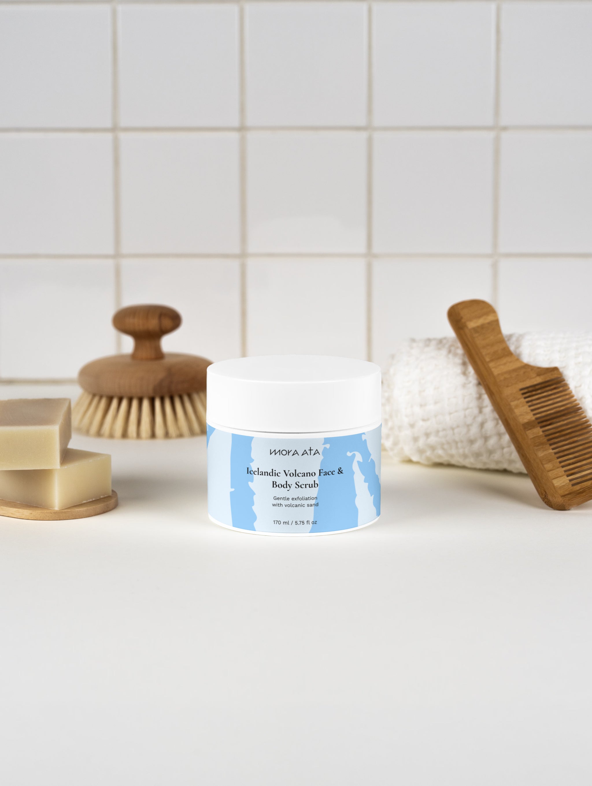 Jar of Mora Ata Icelandic Volcano Face & Body Scrub with a brush, soap, and towel on a tiled bathroom counter