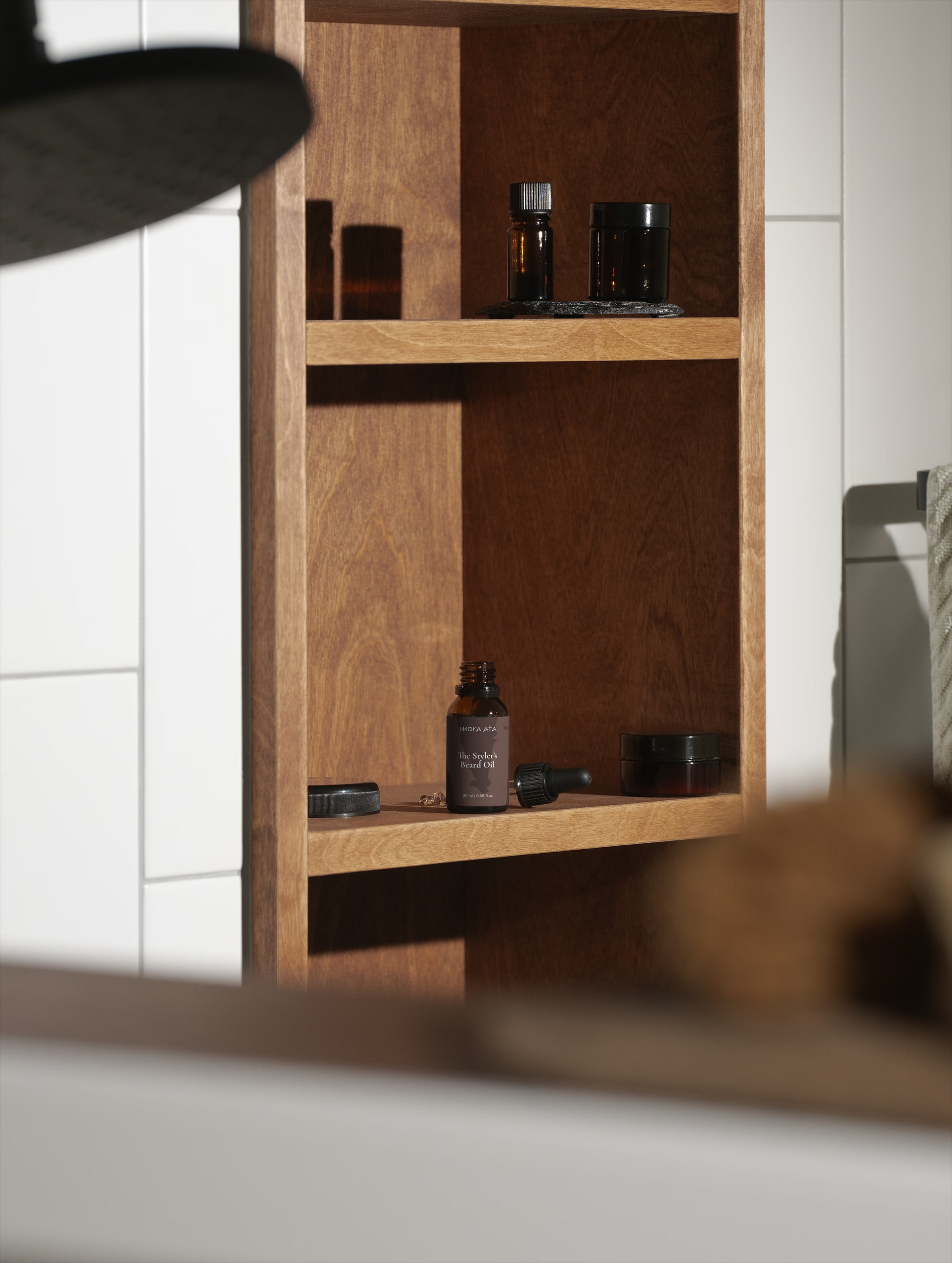 Wooden shelf with skincare products against a white tiled wall featuring a Mora Ata The Styler's Beard Oil bottle with its dropper.
